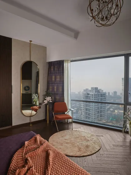 A view of the dark-themed master bedroom, showcasing the cozy reading corner with a view of the Mumbai skyline, herringbone wood flooring, and a dramatic hanging mirror.