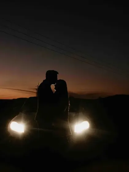 A dramatic silhouette of the couple kissing in front of the car's headlights at dusk, creating a powerful and romantic image.