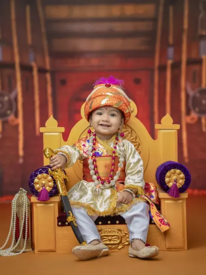 A regal portrait of a baby boy in a Bal Shivba costume, holding a toy sword. The rich fabrics of the costume and the ornate throne create a picture of miniature royalty.
