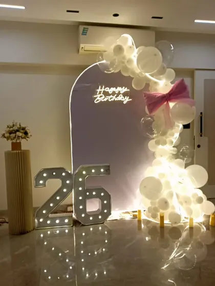 A minimalist and elegant setup for a 26th birthday. A simple arched backdrop with a neon sign is paired with a delicate white balloon garland and large marquee numbers.
