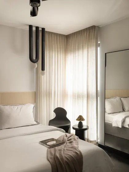 This guest bedroom at Ecru Maison features a minimalist design with a custom fluted headboard panel. A sculptural black chair and a unique U-shaped pendant light add artistic, modern touches to the calm space.