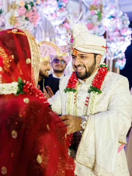 The groom's turn. Aakash smiles as Shruti prepares to place the garland on him during their Mangalashtaka ceremony, with friends and family cheering them on.