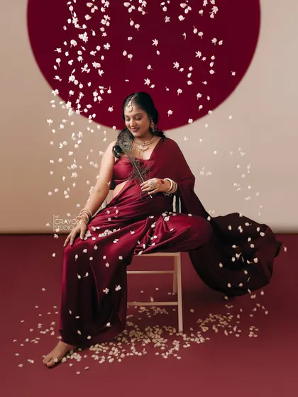Seated gracefully amidst falling petals, she holds a peacock feather, a symbol of divinity and beauty. This artistic studio shot is a celebration of creation.