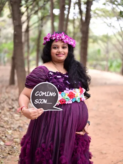 A happy mom-to-be announcing her pregnancy with a 'Coming Soon' sign, wearing our purple ruffle gown in a beautiful forest setting.