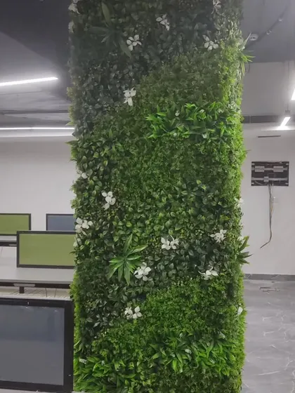 An office pillar wrapped in a dense artificial green panel with white flowers. This is a great way to hide structural elements and bring nature into an open-plan office.