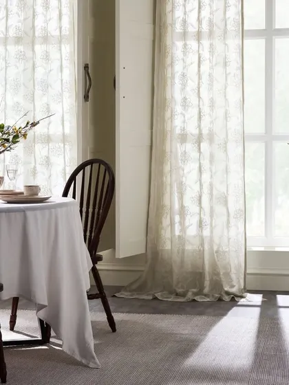 A dining area with light filtering through sheer, embroidered curtains. The image captures the ethereal quality of the fabric and how it interacts with sunlight.