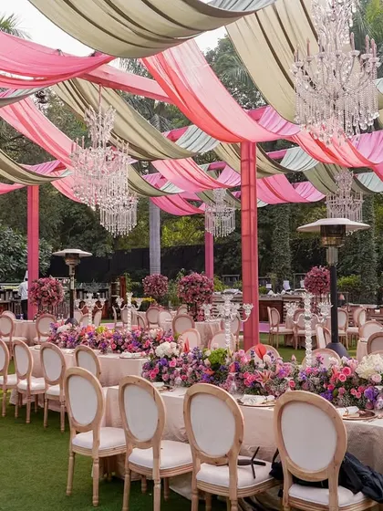 An elegant outdoor dining setup for a garden party. The tables are adorned with lush pink and purple floral runners, under a ceiling of draped fabric and crystal chandeliers.