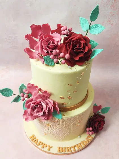 We showcase our artistry with this two-tiered cake featuring handmade edible sugar flowers. Intricate red and pink roses with delicate teal leaves adorn the cake, which also features an elegant embossed pattern on the bottom tier, perfect for a wedding or engagement.