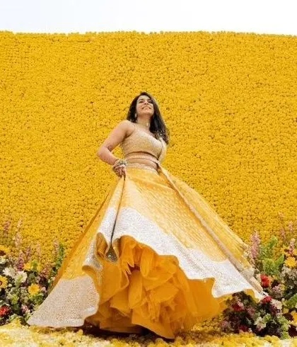 A bride posing against a massive wall of yellow flowers. This makes for a stunning and impactful photo backdrop for a Haldi ceremony.