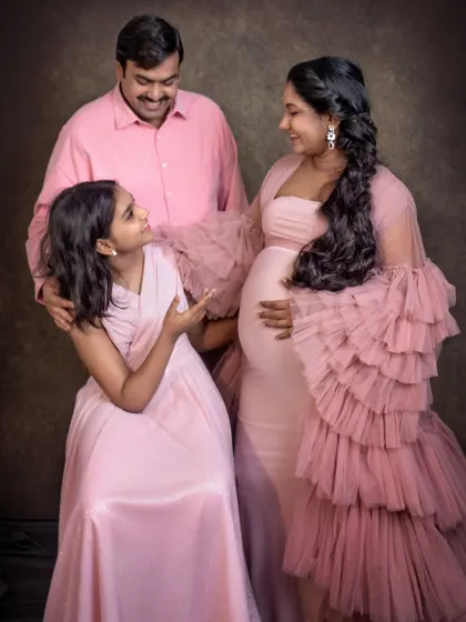 A family of three, all dressed in shades of pink, share a happy moment during a studio maternity shoot. The daughter looks up at her mother, creating a sweet and interactive family portrait.