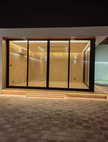 A modern, standalone glass cabin illuminated at night, creating a striking visual. This custom backyard office pod features a black metal frame, warm wood-paneled interiors, and integrated LED lighting along the base, showcasing a premium and contemporary design.