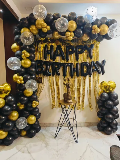 A classic black and gold birthday setup for a home party. This includes a balloon arch over a gold fringe curtain and a small pedestal for the cake.