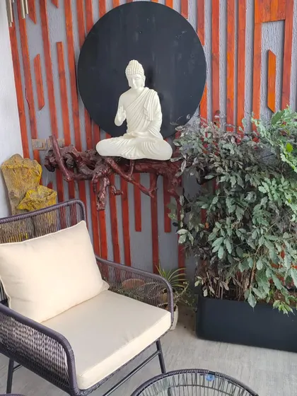 A detailed shot of the Buddha statue on the balcony, placed on a natural wood branch shelf against a textured wooden slat wall. A lush plant in a black planter adds a touch of green to the serene setup.