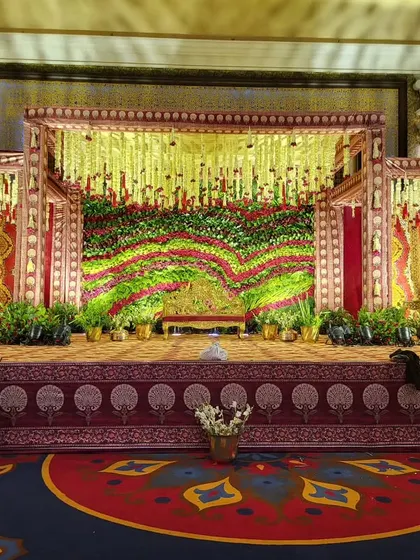 A beautiful example of a traditional stage for a wedding ceremony. The design features a rich floral backdrop using red and green flowers, framed by structures draped in classic wedding colors and patterns.