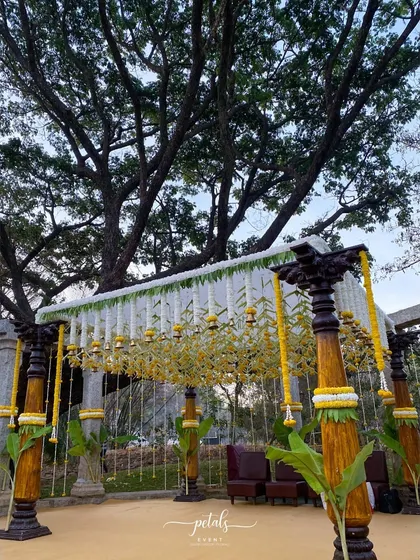The majestic wooden pillars are adorned with white chrysanthemums and temple bells, creating a classic South Indian wedding vibe. The plantain leaves add a final touch of authenticity and cultural charm.