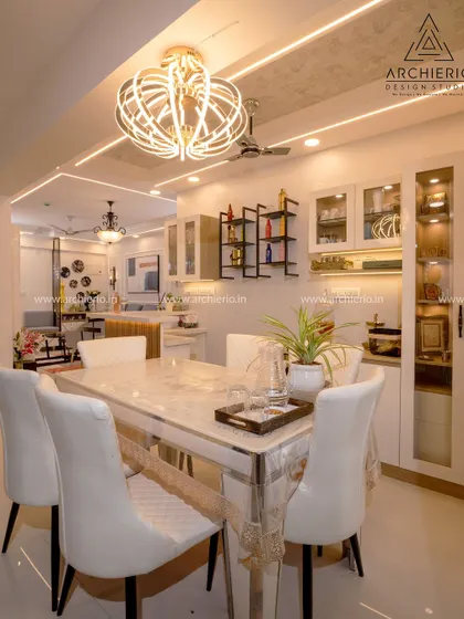 A grand dining room with a marble-topped table and elegant white chairs. The statement chandelier and well-lit crockery unit add to the luxurious feel of the space.