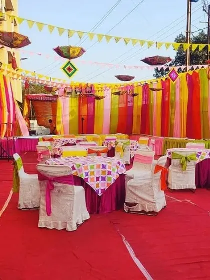 A large outdoor Mehendi event setup with colorful draped walls and hanging umbrellas. The space is arranged with tables and chairs for guest seating.