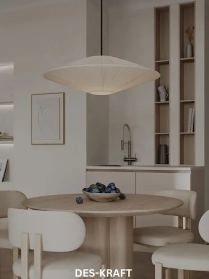 A minimalist dining space featuring a round wooden table, soft upholstered chairs, and a large, sculptural pendant light, embodying the Japandi style.