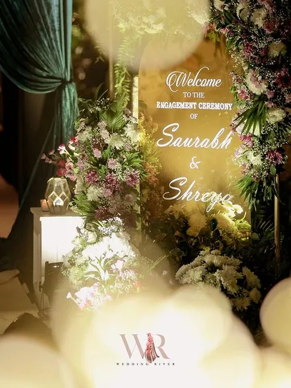 A soft, romantic welcome for Saurabh and Shreya's engagement. The custom-lit sign is nestled within an arrangement of pink and white florals, with the bokeh of surrounding lights creating a dreamy, ethereal effect.