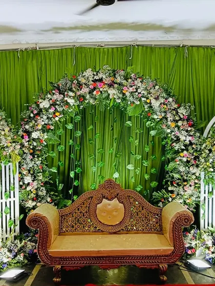A lush and green engagement stage. The backdrop features a large floral arch with hanging leaves, flanked by smaller white arches, creating a beautiful garden-like setting.