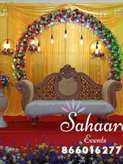 A vibrant reception stage with a floral arch against a bright yellow backdrop. The hanging lanterns and ornate sofa create a festive and traditional setting for any celebration.
