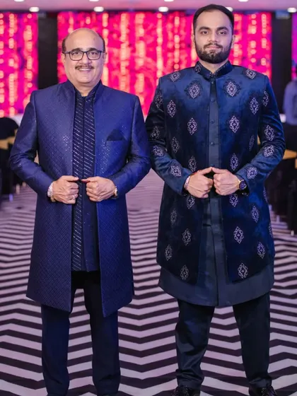 A "like father, like son" moment, with both wearing custom blue Indo-Western outfits. The son's jacket features velvet appliqué and embroidery, while the father's is a classic brocade Jodhpuri.