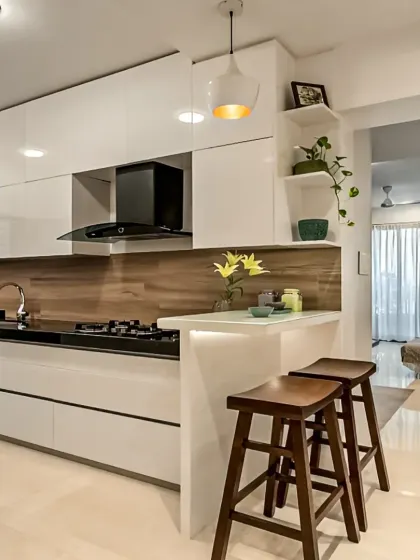 This open kitchen design includes a breakfast counter with stylish wooden stools, perfect for quick meals. The all-white, high-gloss cabinets make the space feel bright and spacious, connecting seamlessly with the living area.