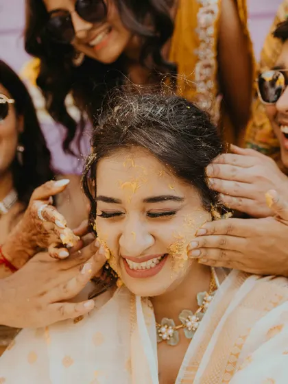 The bride closes her eyes and smiles as her friends and family lovingly apply Haldi paste. A beautiful, candid moment of tradition and affection.