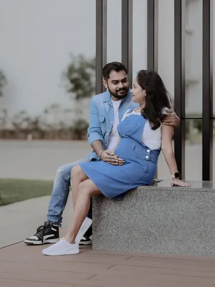 A candid and happy moment between an expecting couple during their modern maternity shoot. Dressed in casual denim, they are seated outdoors, sharing a look of affection.