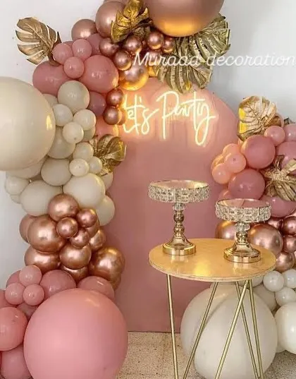 A vibrant and fun party corner with a "Let's Party" neon sign. The balloon garland in shades of pink, cream, and rose gold is accented with gold-painted tropical leaves, creating a stylish setup for the cake table.