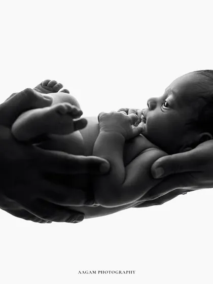 A creative black and white newborn portrait, showing the baby held horizontally in their parents' hands. The silhouette effect is striking and artistic.