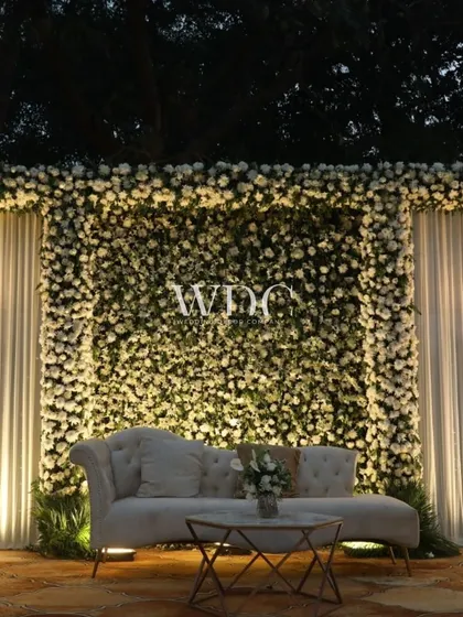 A classic and timeless stage setup. A wall of white flowers serves as the perfect backdrop for an elegant ivory-colored lounge sofa, creating a look of pure sophistication.