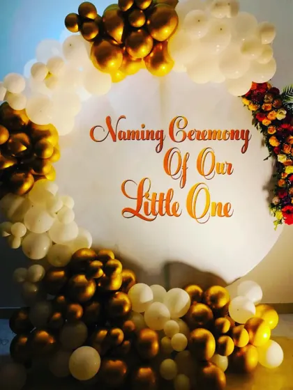 An elegant naming ceremony backdrop with a simple and clean design. The focus is on the "Naming Ceremony Of Our Little One" text, framed by a beautiful arch of gold and white balloons with a touch of floral arrangement.