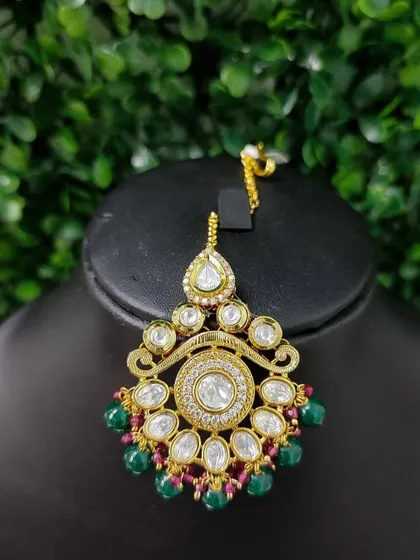 A kundan maang tikka with a central clear stone, surrounded by smaller stones and a mix of red and green bead drops.