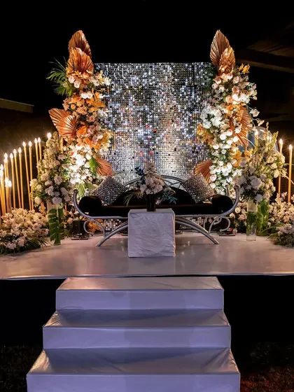 A stunning stage setup for an engagement or Sangeet, featuring a shimmering silver sequin backdrop, lush florals, and elegant candle lighting.
