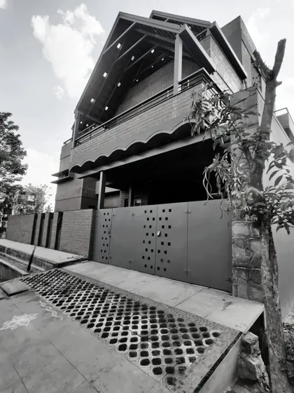 A black and white photograph of the Madhura residence, highlighting its strong architectural form, textures, and the dramatic sloping roofline.