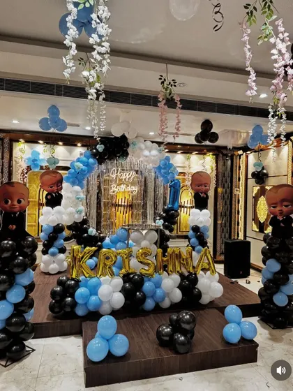 A stage decoration combining a Boss Baby theme with hanging floral elements. The setup includes character cutouts on balloon pillars and the name 'Krishna' in foil balloons.