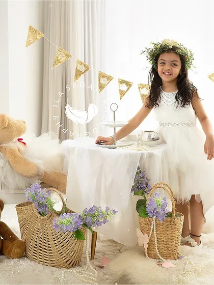 A full view of the whimsical tea party setup. This toddler is standing by the table, ready to host her party in a beautiful white dress.