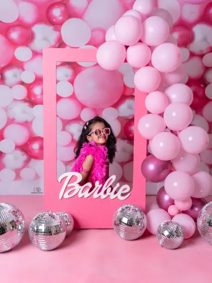 A vibrant 'Barbie' themed photoshoot for a stylish toddler. She poses confidently in a pink frame against a wall of pink balloons and disco balls, complete with a feather boa and sunglasses.
