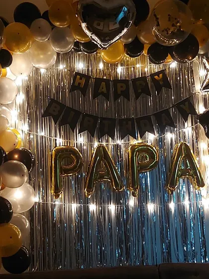 A special birthday decoration for "PAPA". The setup includes a silver foil curtain with fairy lights, a balloon arch, and gold foil letters, creating a heartfelt surprise.