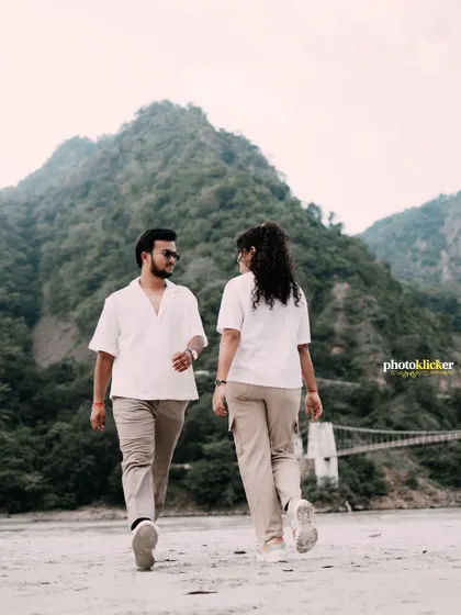 A candid walking shot of a couple on a riverbank, with a suspension bridge visible in the background, capturing a natural interaction.