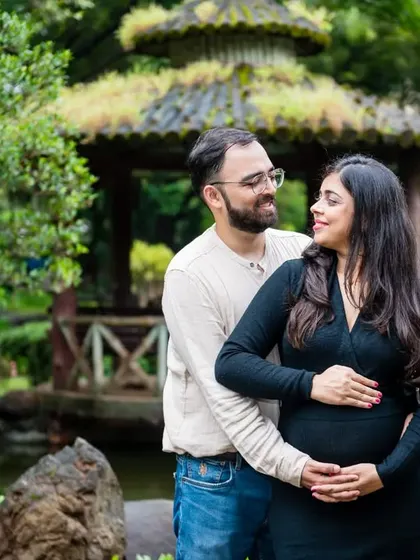 A classic pose with the father-to-be standing behind, cradling the baby bump. The serene park setting with the gazebo in the background adds a romantic touch to this maternity photo.