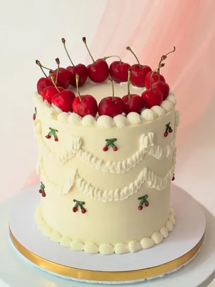 A charming vintage-style cake with classic white swag piping and tiny piped cherries. The cake is crowned with a ring of fresh, glossy cherries.