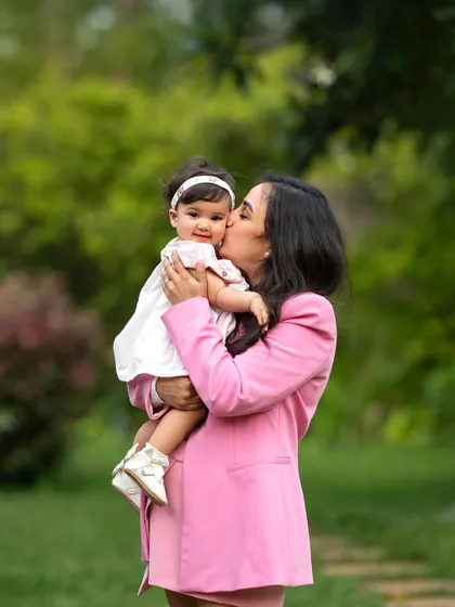 A mother in a stylish pink suit holds her baby girl, who is smiling sweetly. These mother-daughter portraits in the park are always full of love.