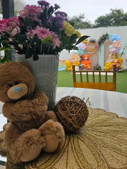 A close-up of a table centerpiece at a teddy bear-themed baby shower. A cute teddy bear sits next to a vase of flowers, with the main party setup visible in the background.