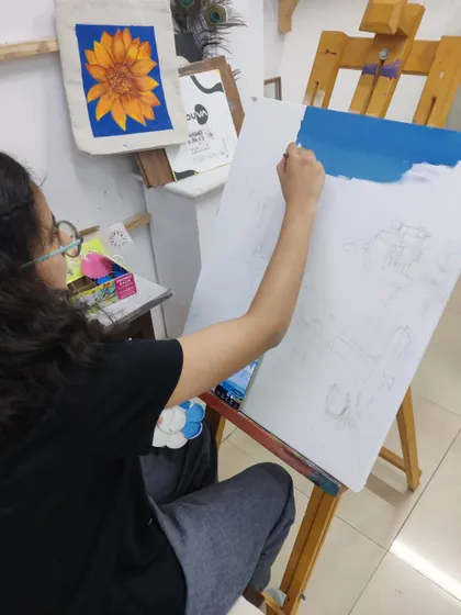A student begins her Santorini landscape painting, sketching the composition on canvas and applying the first layer of blue for the sky.
