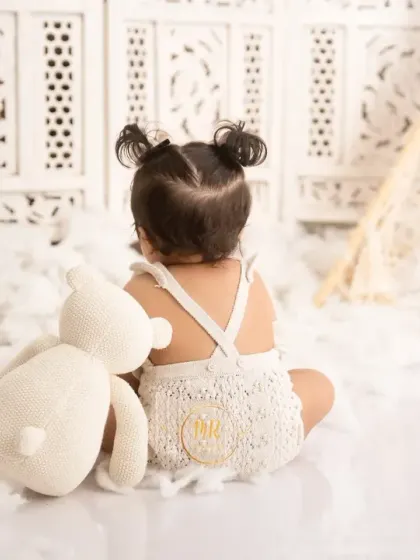 A quiet moment from behind, showing a baby girl with her teddy bear, highlighting her cute pigtails and knitted romper.
