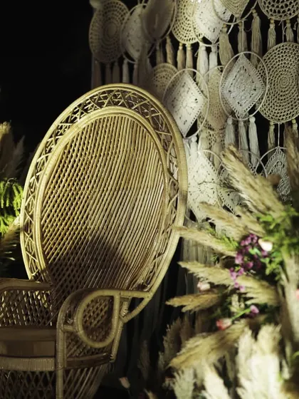 A bohemian-themed Mehendi night detail, showing a peacock chair against a macrame backdrop, surrounded by pampas grass.