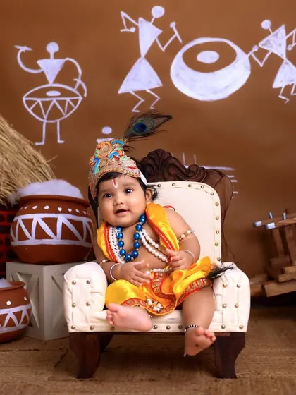 A smiling little Krishna seated on his throne. The composition with the surrounding matkis and village backdrop creates a complete and festive picture.