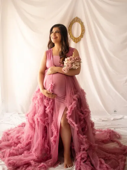 A radiant solo portrait of a mother-to-be in a pink gown, looking up with a joyful expression. The simple white backdrop keeps the focus on her.